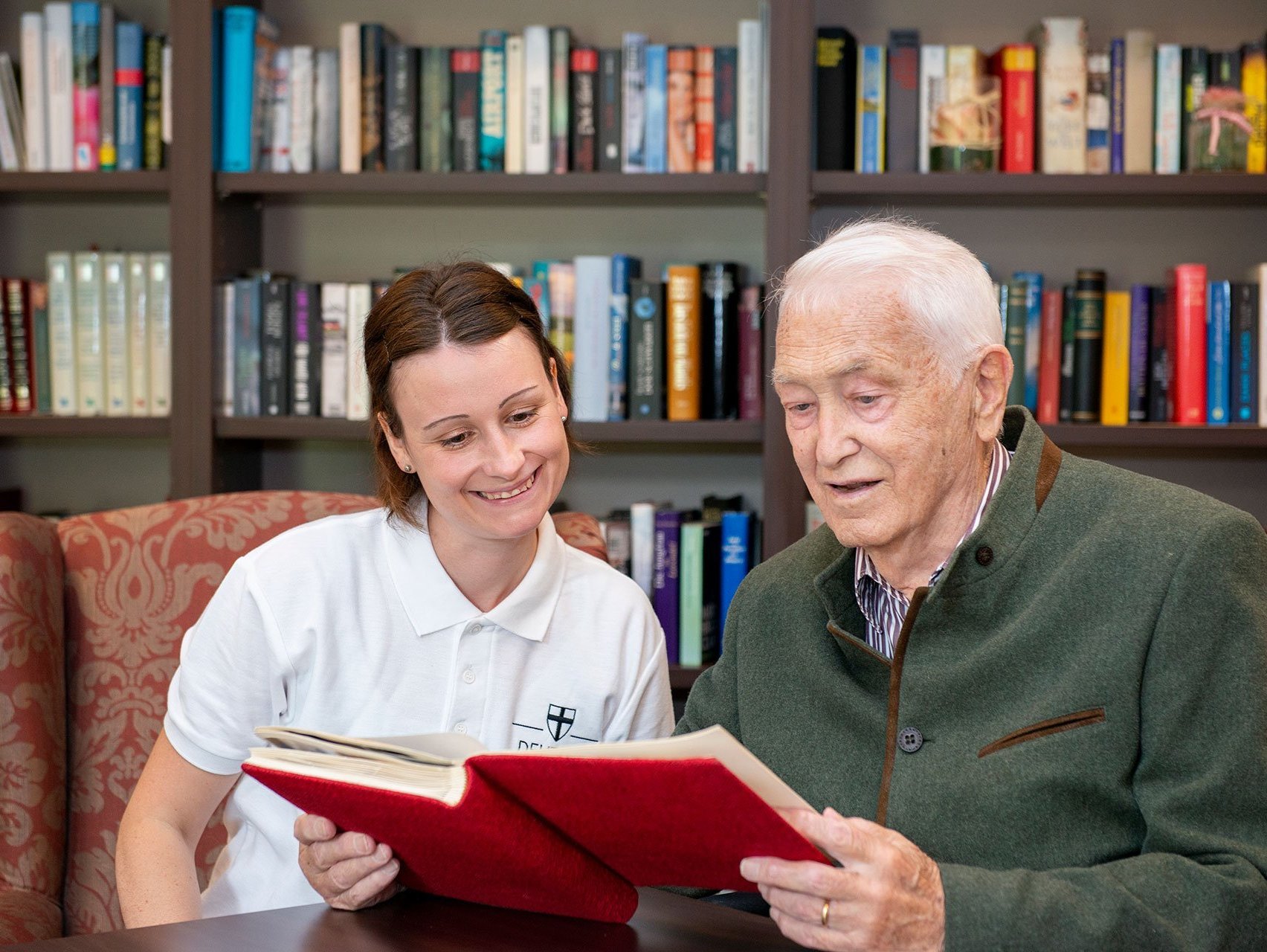 Ein älterer Mann liest aus einem Buch während eine Pflegerin zusieht
